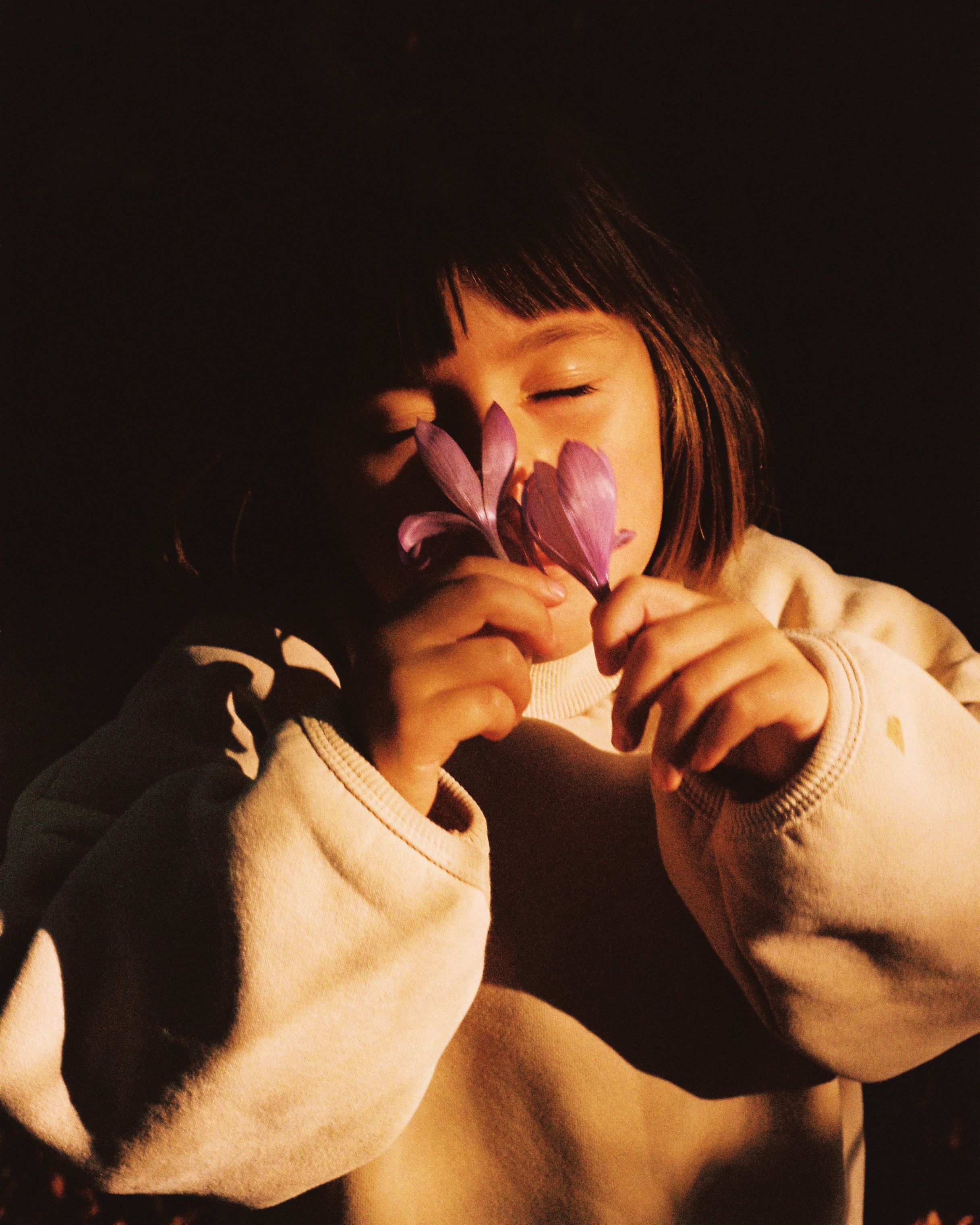 A young girl with short hair and a white sweater is holding a purple flower and smiling.