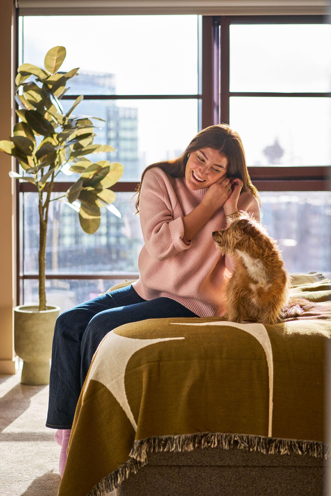 A woman in a pink sweater and jeans sitting on a bed with a small dog on her lap, in a room with a city skyline visible through the window.