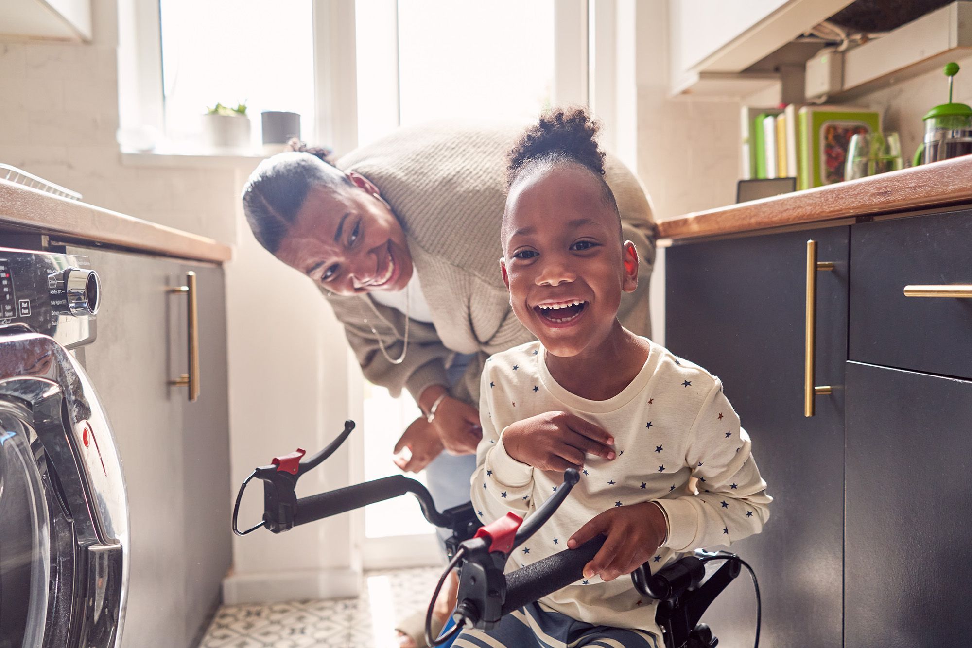 A woman in a kitchen with a young girl on a bicycle, with a washing machine and kitchen cabinets in the background.