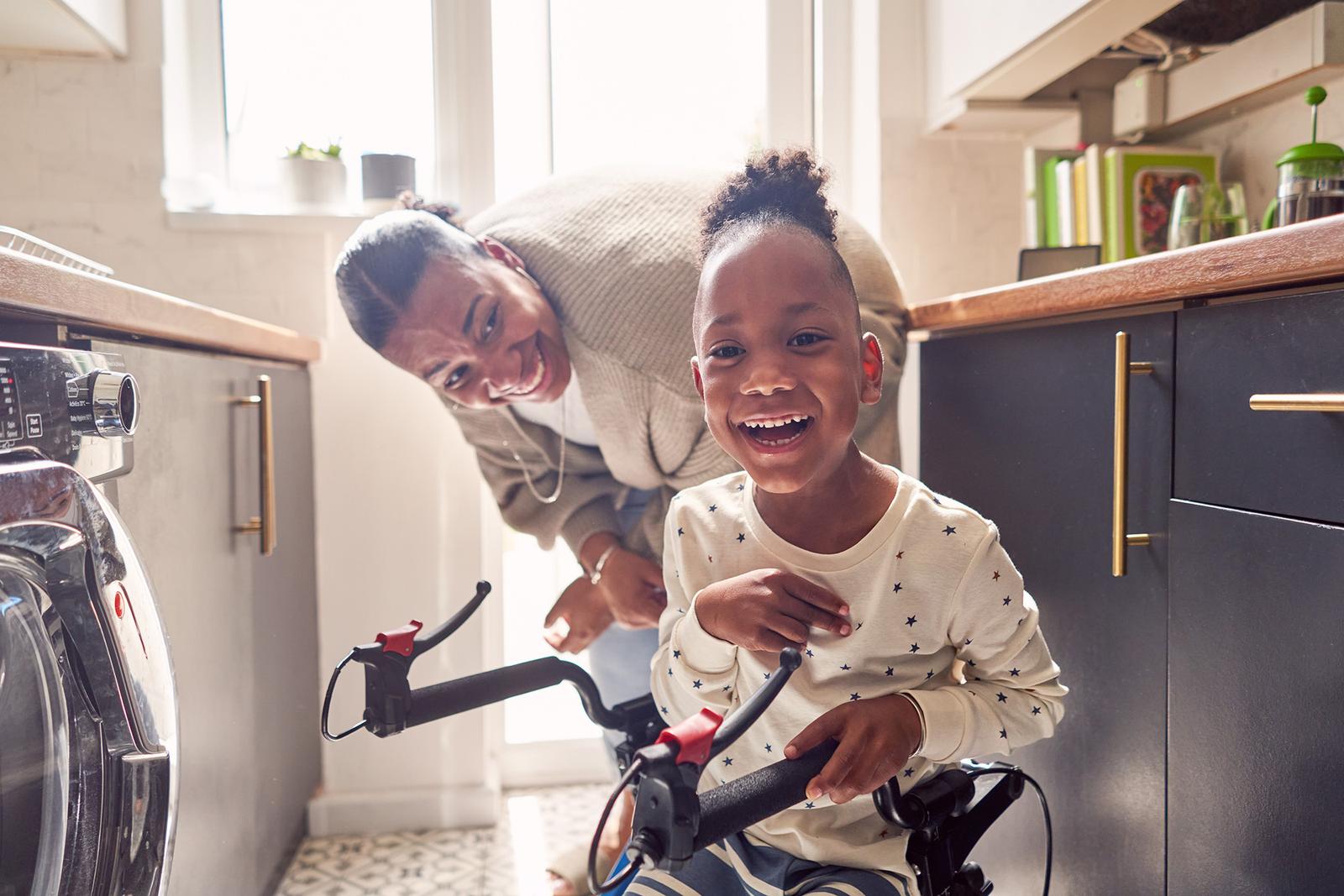 A woman in a kitchen with a young girl on a bicycle, with a washing machine and kitchen cabinets in the background.