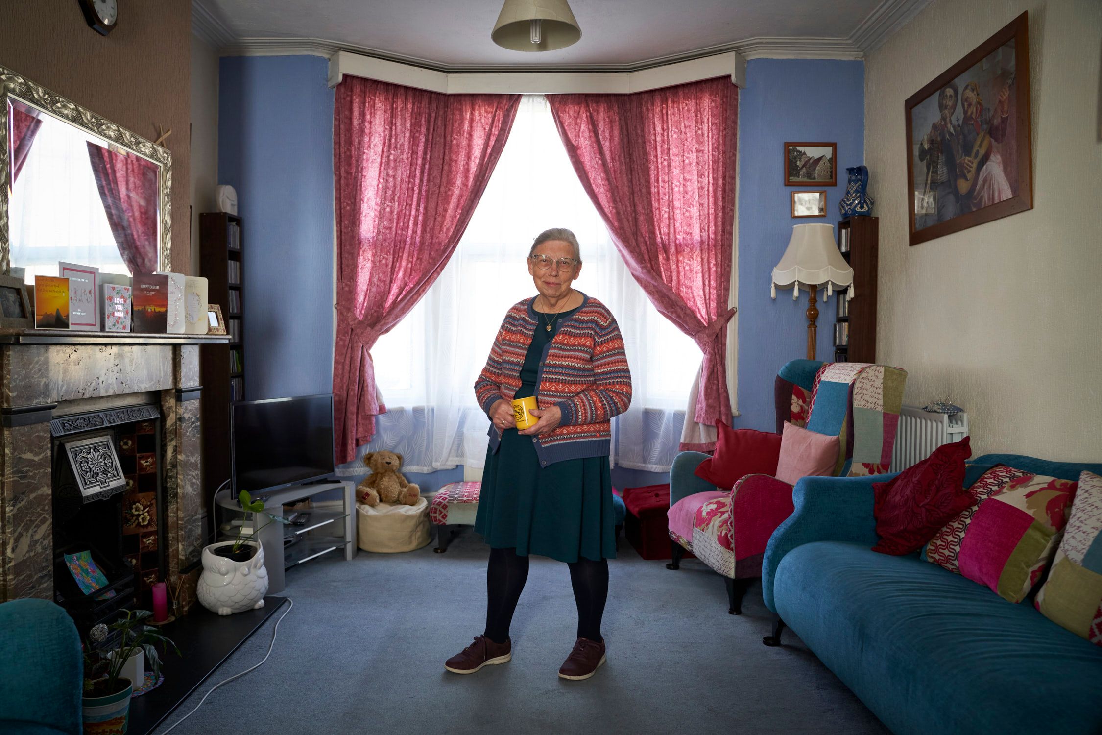 An elderly woman stands in a living room, wearing a striped cardigan, a blue dress, and holding a yellow cup.