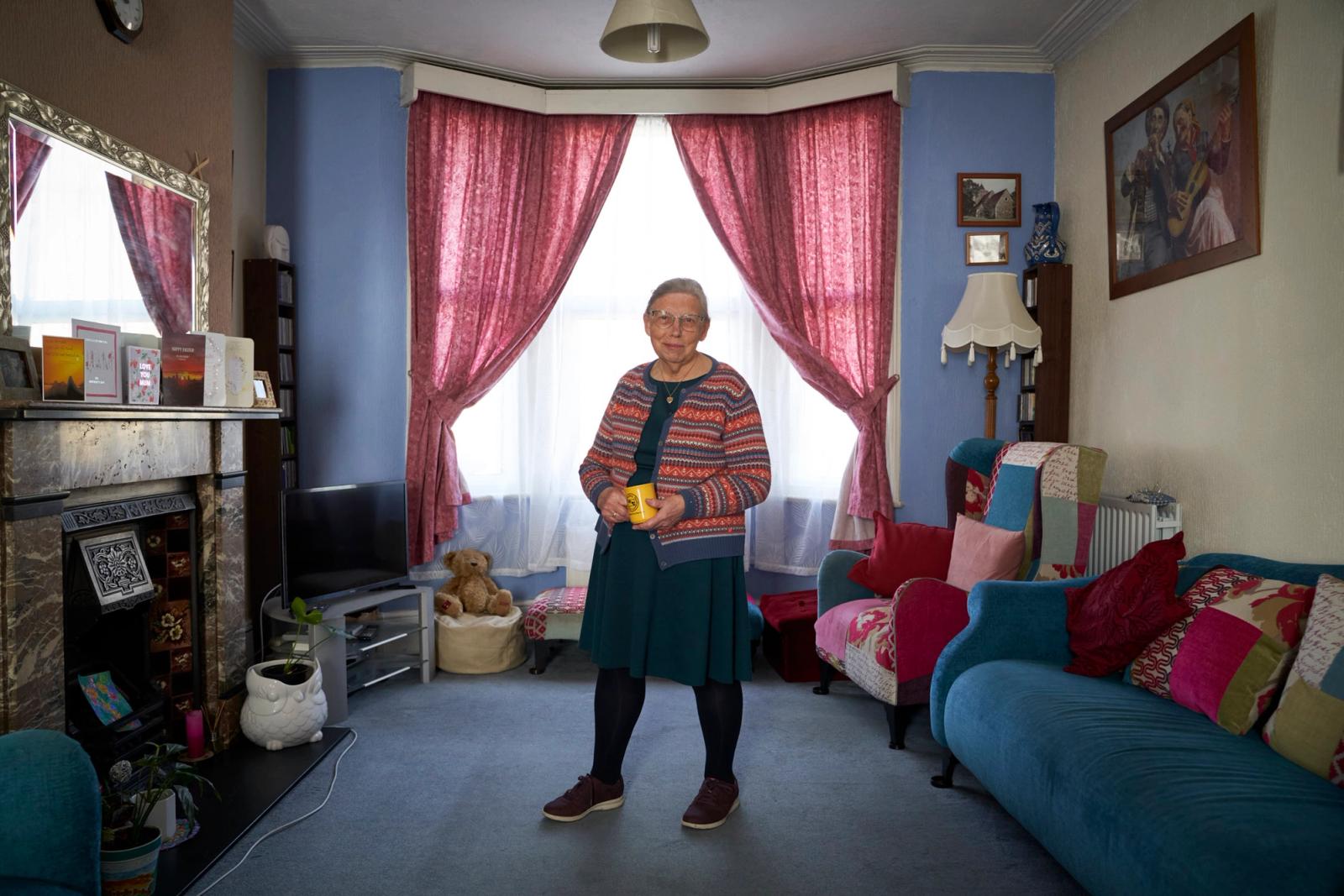 An elderly woman stands in a living room, wearing a striped cardigan, a blue dress, and holding a yellow cup.