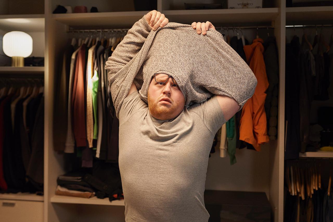 A man in a grey t-shirt is standing in front of a closet, holding up a grey sweater with both hands, covering his head.