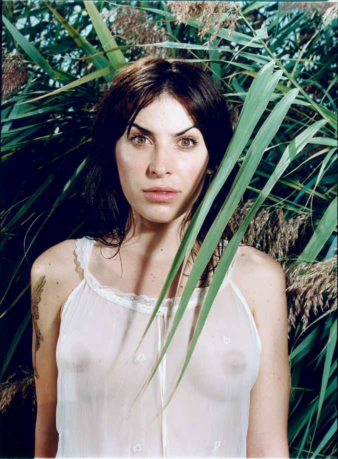 A woman with dark hair and a white tank top standing in front of a green plant with long, thin leaves.
