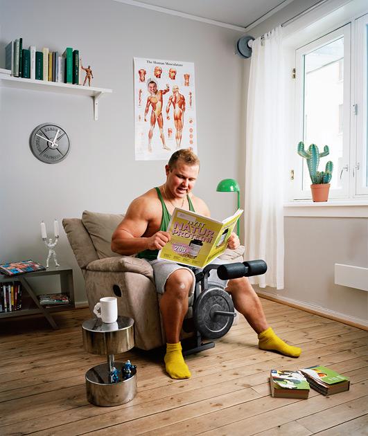 A man in a green tank top and grey shorts is sitting in a chair, reading a magazine called 'Morten Borgestad Portfolio 2019'.