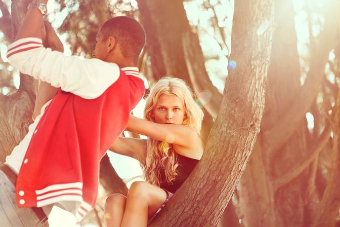 A young woman with long blonde hair and a black tank top is sitting on a tree branch, with a young man in a red and white striped rugby jersey standing next to her, both looking directly at the camera.