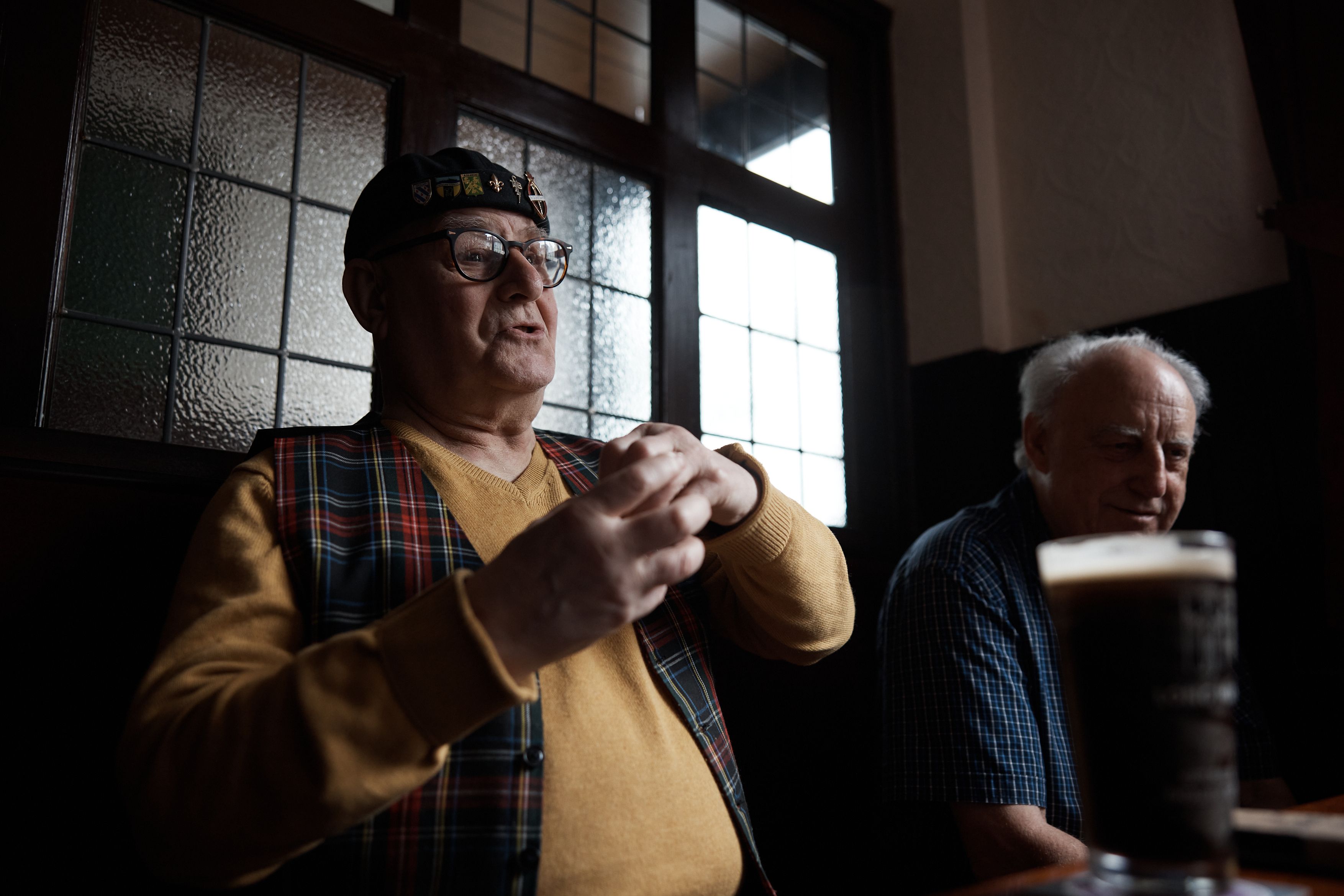 Two older men sit at a table in a dimly lit pub, one dressed in a tartan waistcoat and black cap with a yellow and red logo, the other in a blue and white plaid shirt.