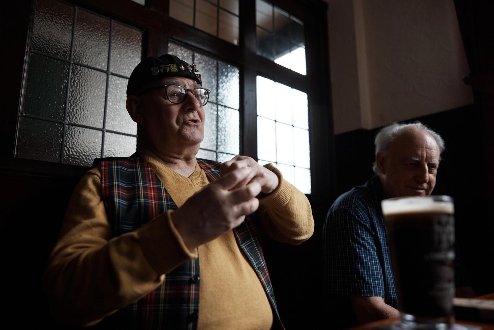 Two older men sit at a table in a dimly lit pub, one dressed in a tartan waistcoat and black cap with a yellow and red logo, the other in a blue and white plaid shirt.