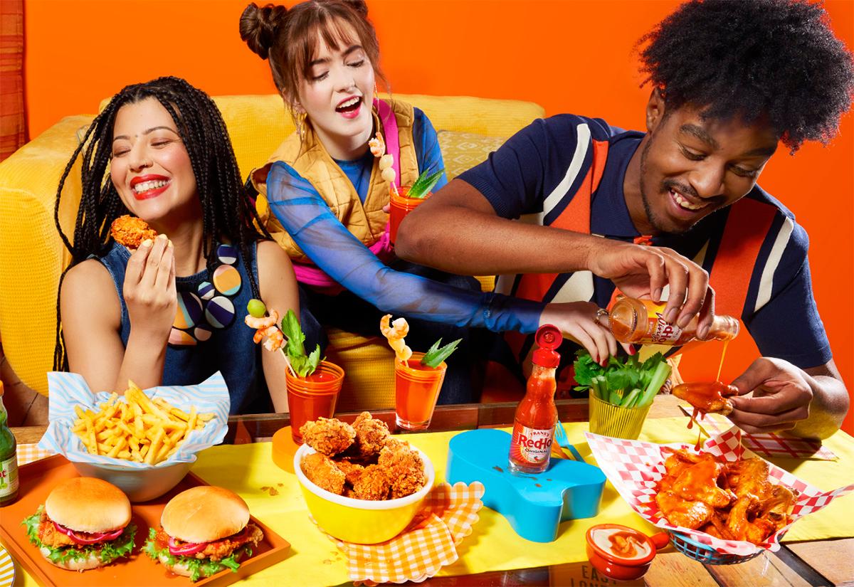 A group of people, including a woman with long, dark hair and a man with a beard, are sitting at a table with a variety of food and drinks, including burgers, fries, and cocktails.