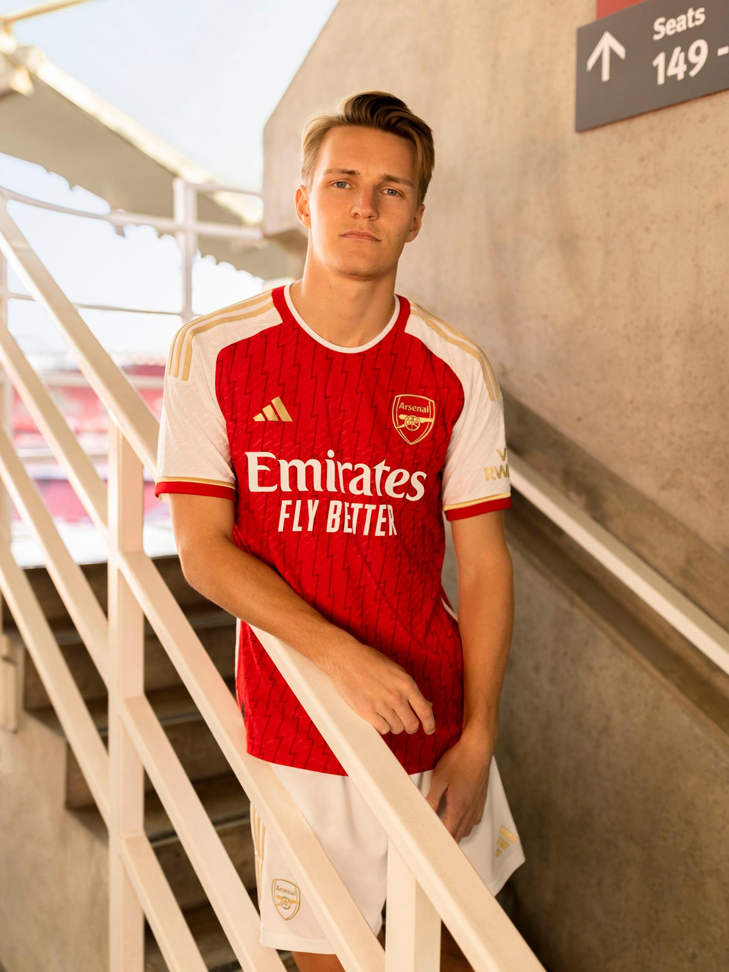 A young man wearing a red and white football jersey with the Arsenal logo on it, standing in front of a concrete wall with a sign pointing to the stadium's seats.