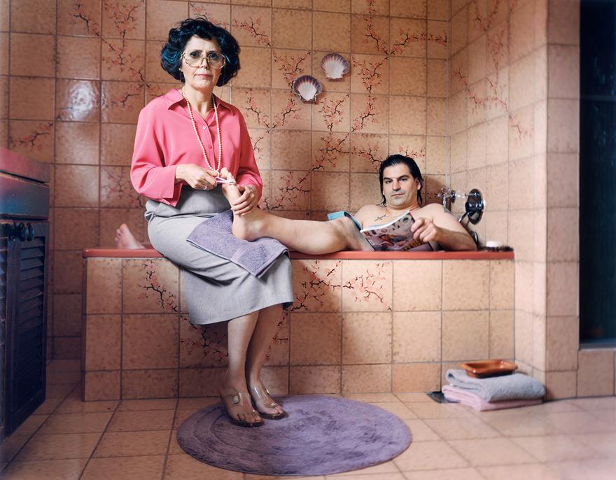 A woman in a pink blouse and grey skirt is sitting on a bench in a bathroom, holding a man's foot and giving him a pedicure.