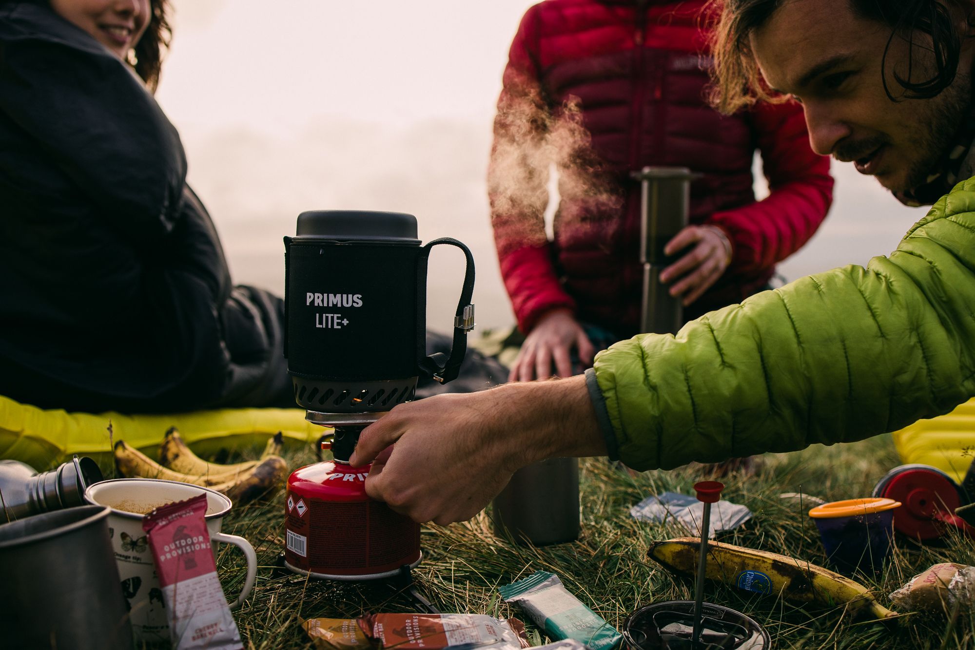 A man in a green jacket is cooking on a portable camping stove in a grassy field, surrounded by camping equipment and food.