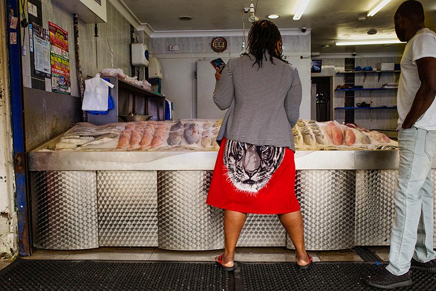 A woman in a red skirt with a tiger print, a striped jacket, and flip-flops is standing in front of a fish counter in a market or store, holding a phone and looking at the fish.