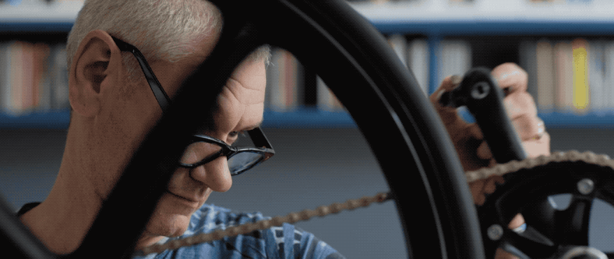 A man with short, white hair and black-framed glasses is working on a bicycle chain in a workshop or library setting, with a bookshelf in the background.