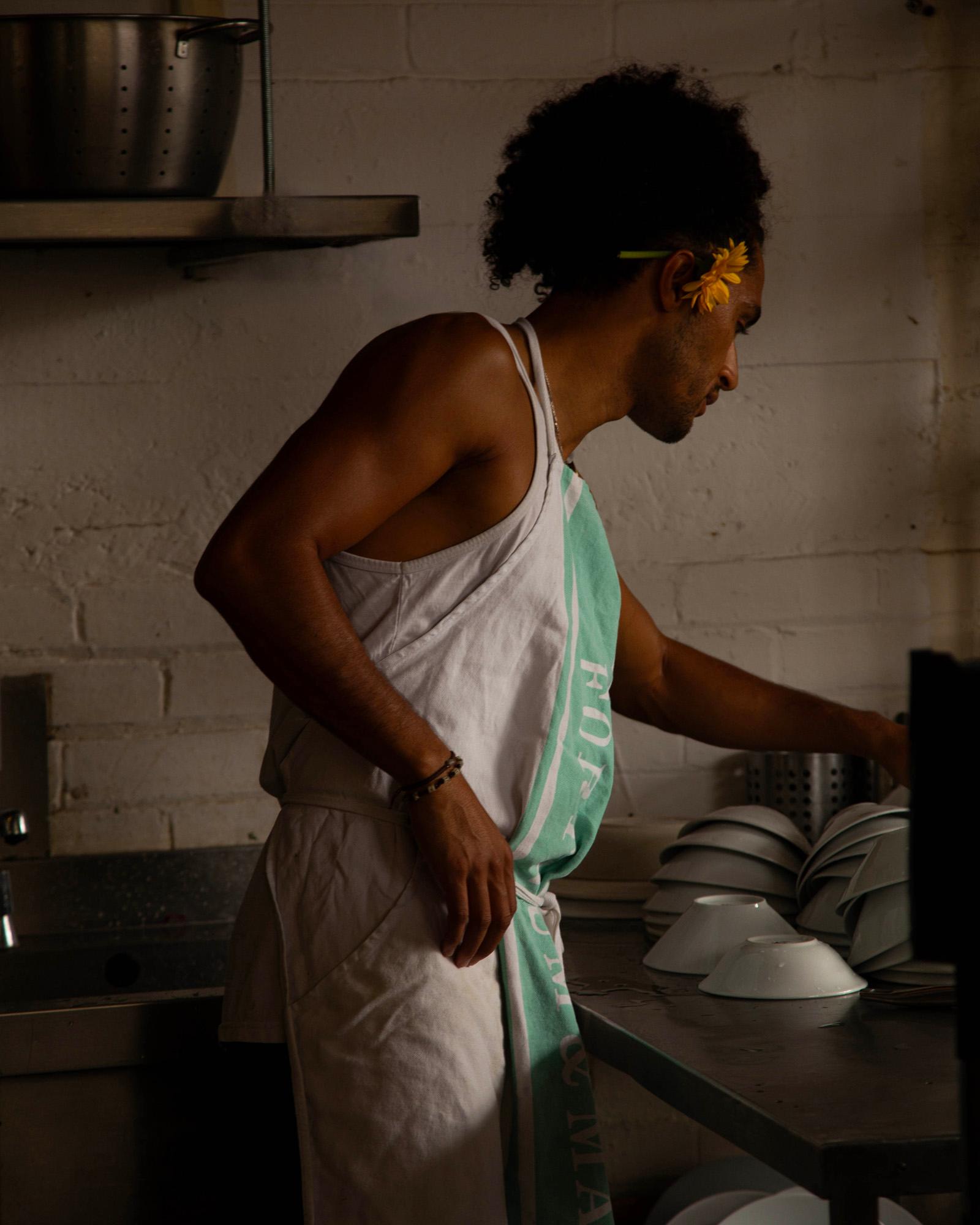 A man in a white apron, with a chef's hat and a flower in his hair, standing in a kitchen, surrounded by cooking utensils and ingredients.
