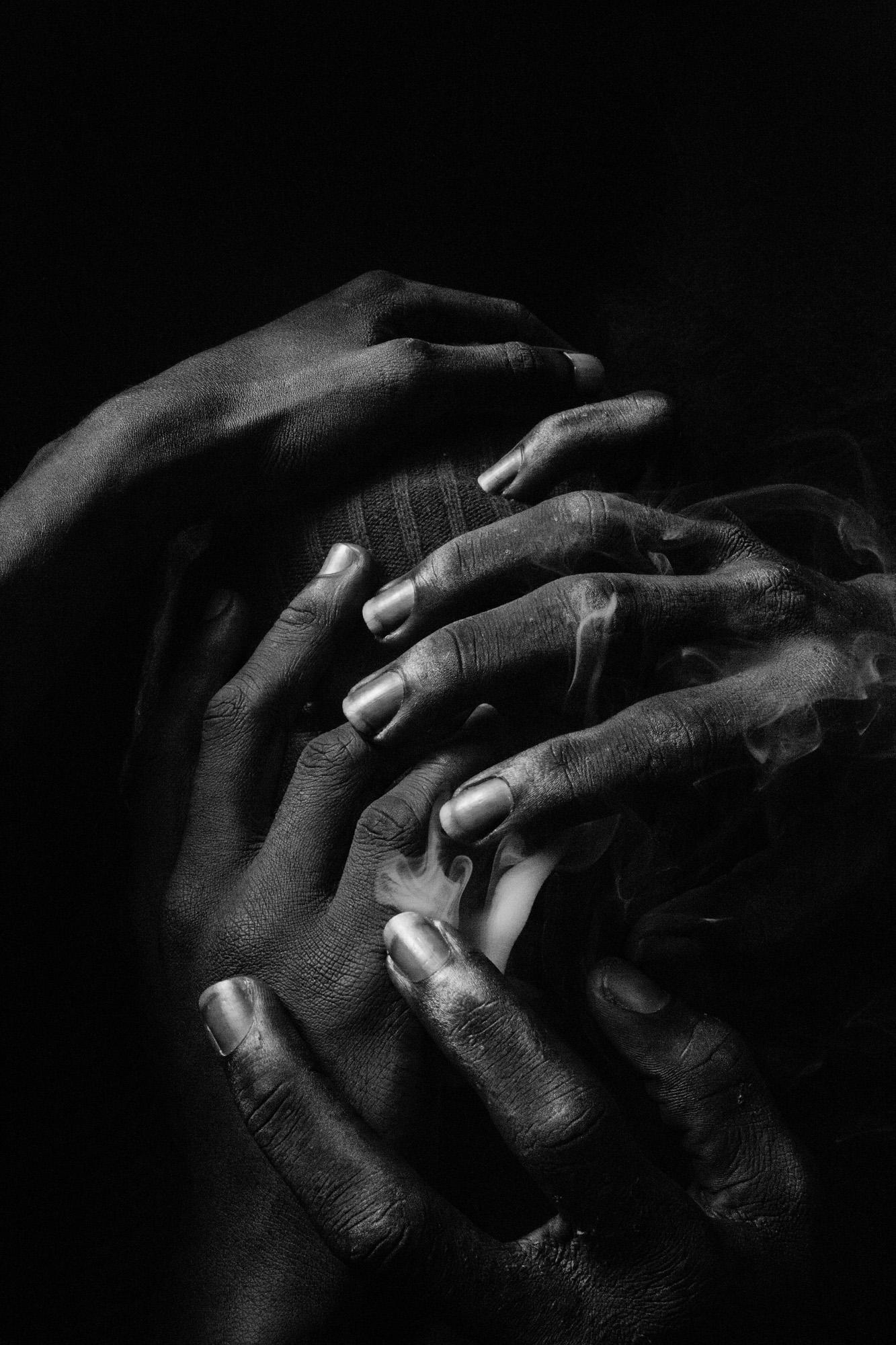 A group of hands, some with fingers intertwined, others with fingers spread, all with dark skin, in a dark, moody, and intimate setting.