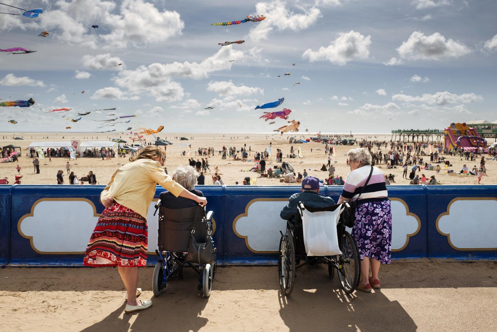 A group of elderly women, one in a wheelchair, watching a beach festival with many colorful kites and people in the background.