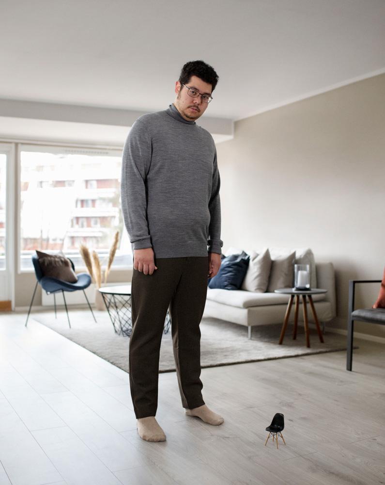 A man in a grey sweater and dark trousers stands in a living room, looking down at a small chair on the floor.