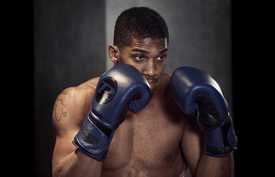 A shirtless, muscular man wearing boxing gloves with a determined expression in a dimly lit room.