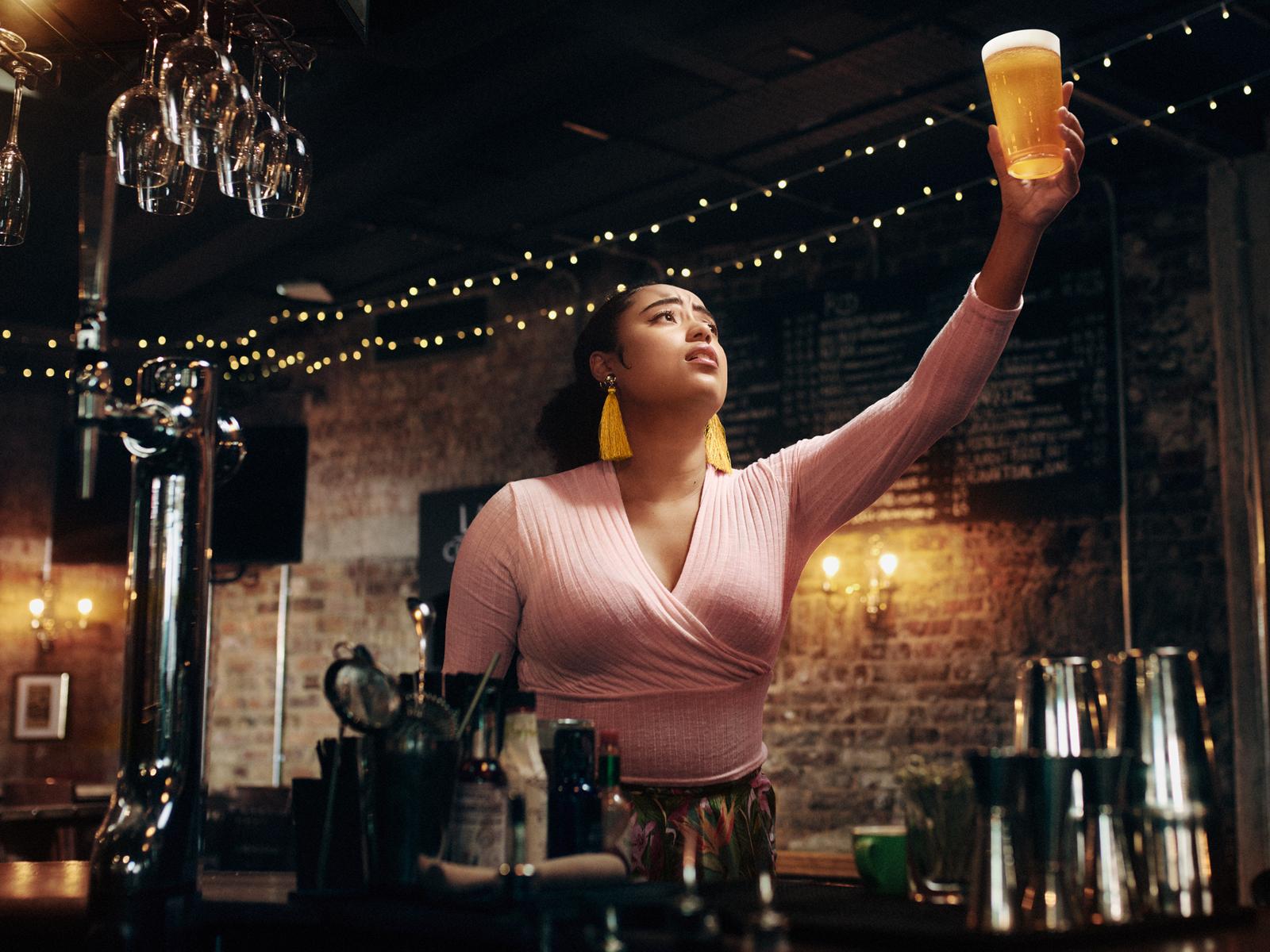 A woman in a pink top and yellow earrings holding a glass of beer in a dimly lit bar.