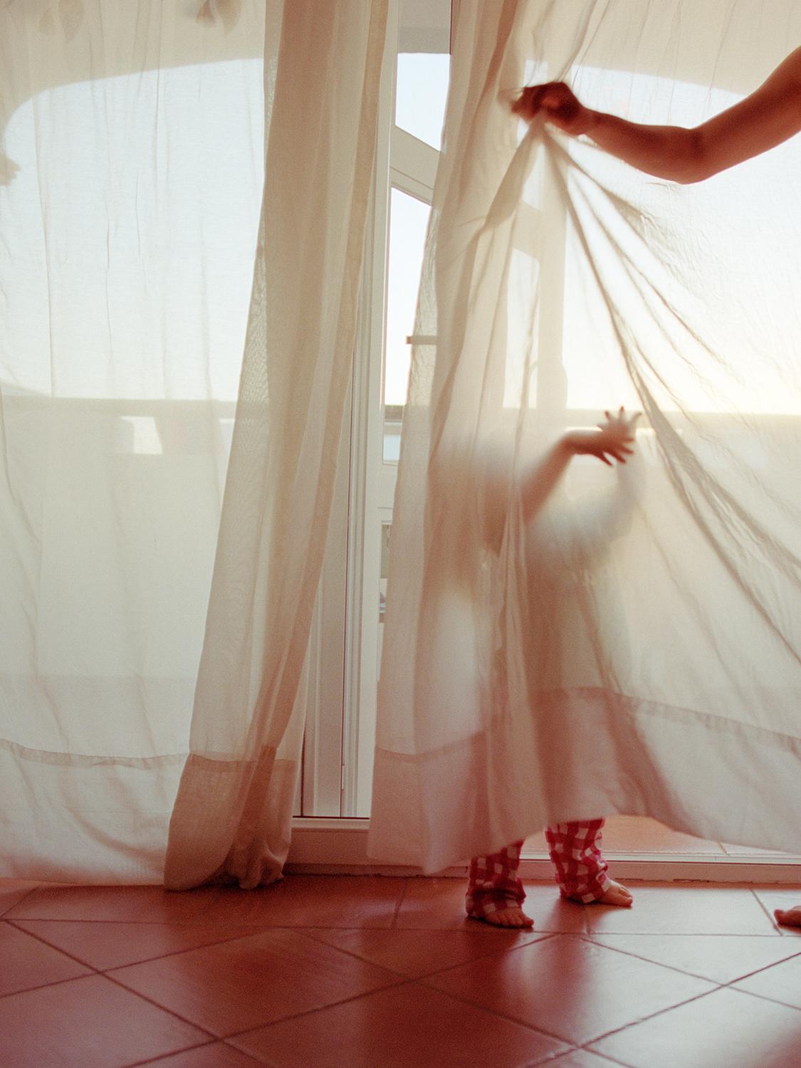 A woman in a white dress is hiding behind a white curtain, with a child's hand and a woman's arm visible through the fabric.
