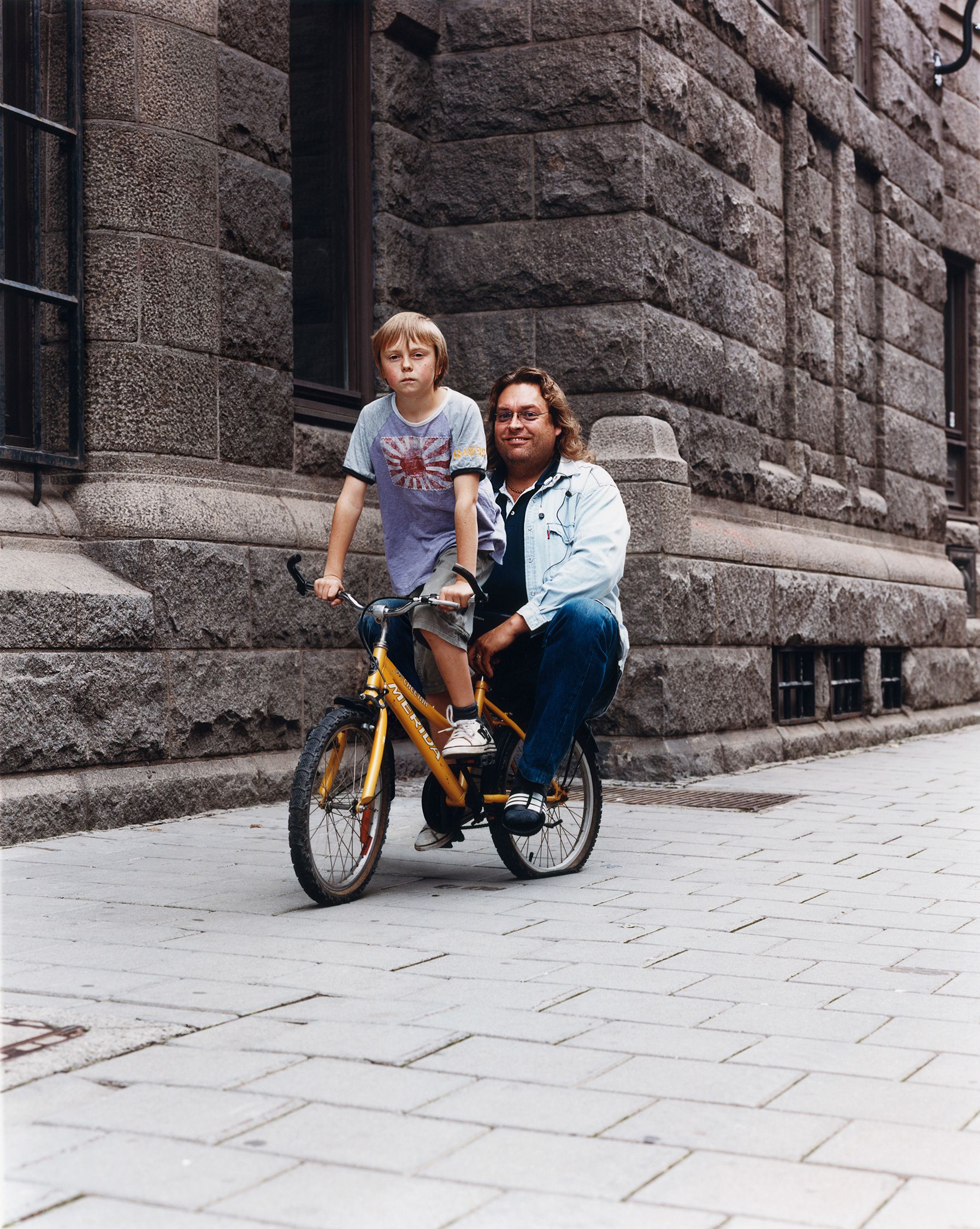 A man and a young boy are riding a yellow bicycle on a stone-paved street in front of a stone building.