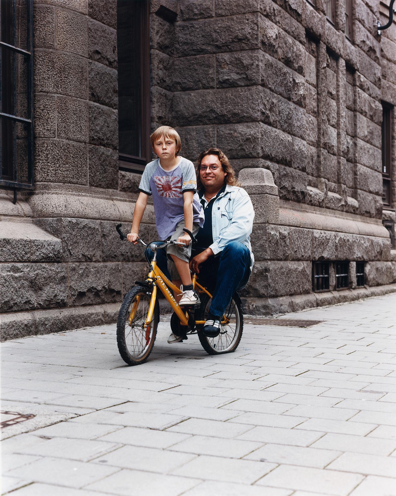 A man and a young boy are riding a yellow bicycle on a stone-paved street in front of a stone building.