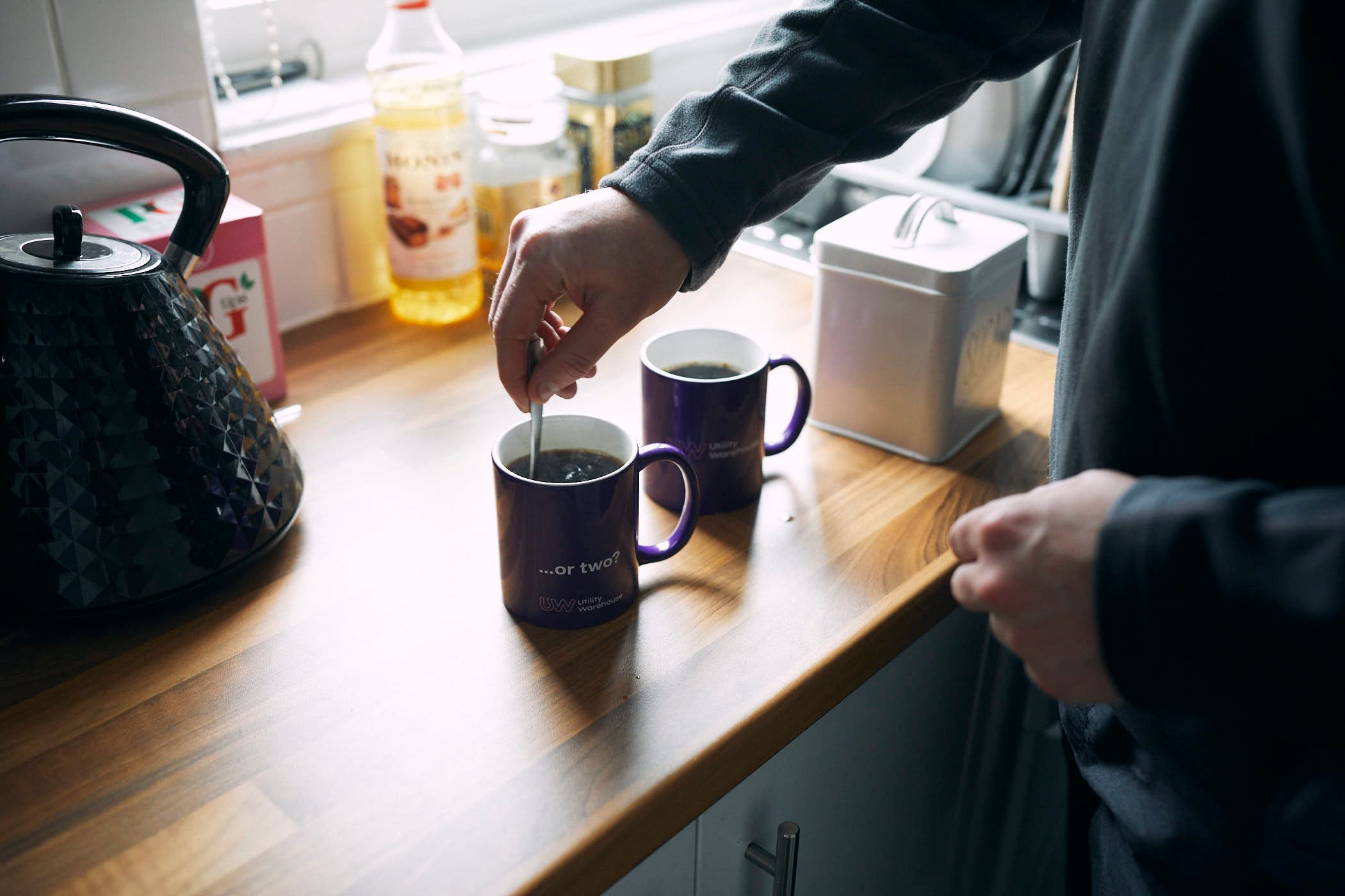 A person stands in a kitchen, stirring a coffee or tea in a purple mug with a white interior and handle. The person is wearing a dark grey long-sleeved shirt and has their arm.