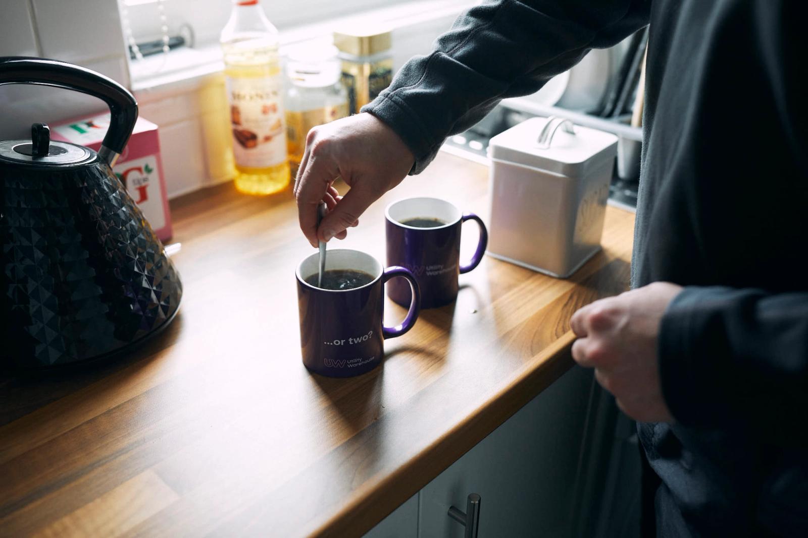 A person stands in a kitchen, stirring a coffee or tea in a purple mug with a white interior and handle. The person is wearing a dark grey long-sleeved shirt and has their arm.