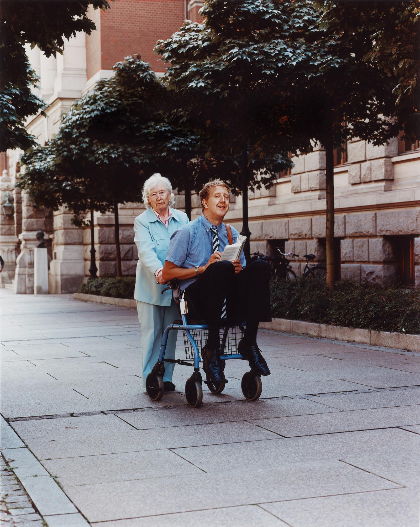 A man in a blue shirt and tie is pushing a shopping cart with a woman in a blue suit standing behind him, both in a city street.