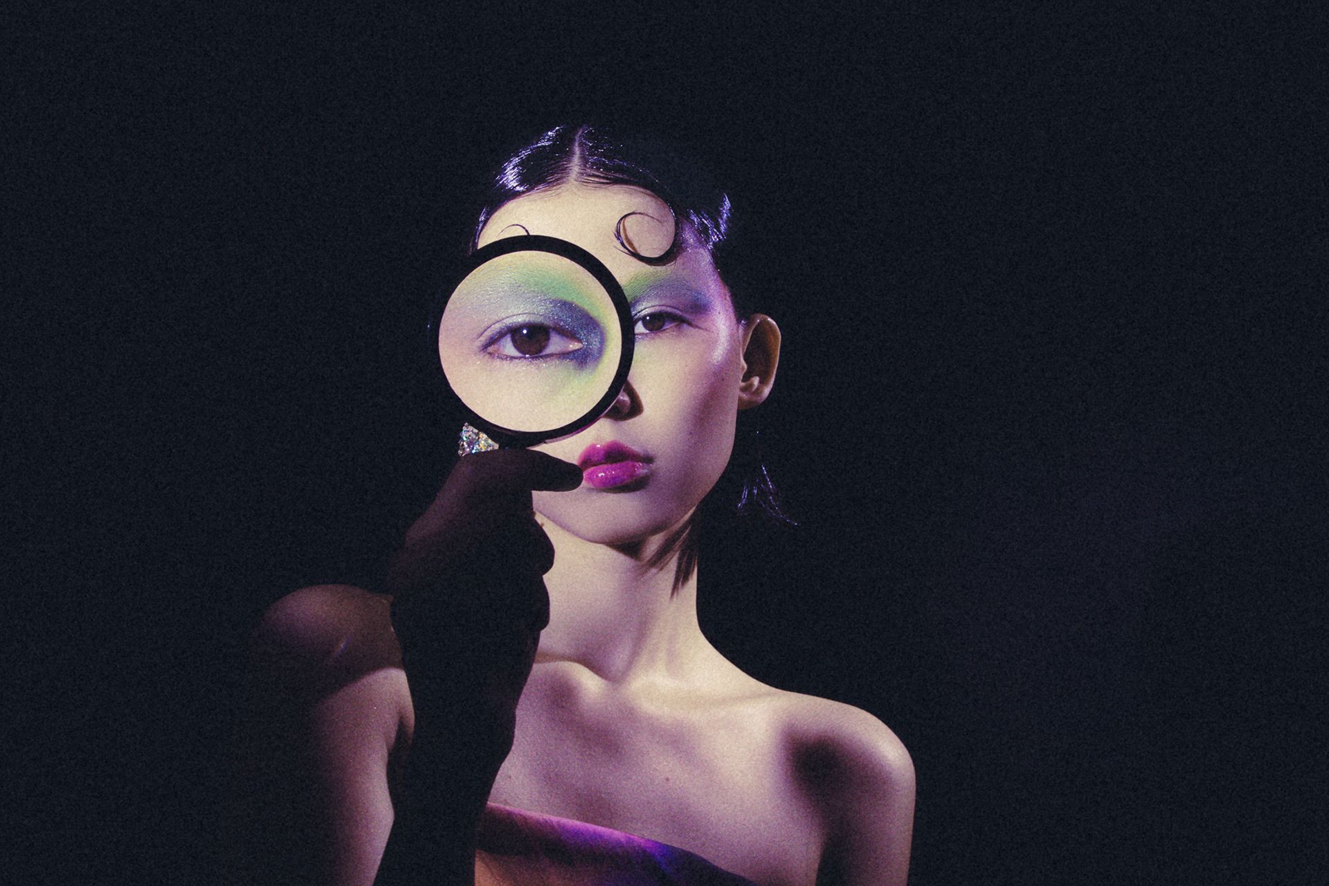 A close-up of a woman's eye, with a magnifying glass held up to it, revealing a vibrant, colorful eye shadow design.