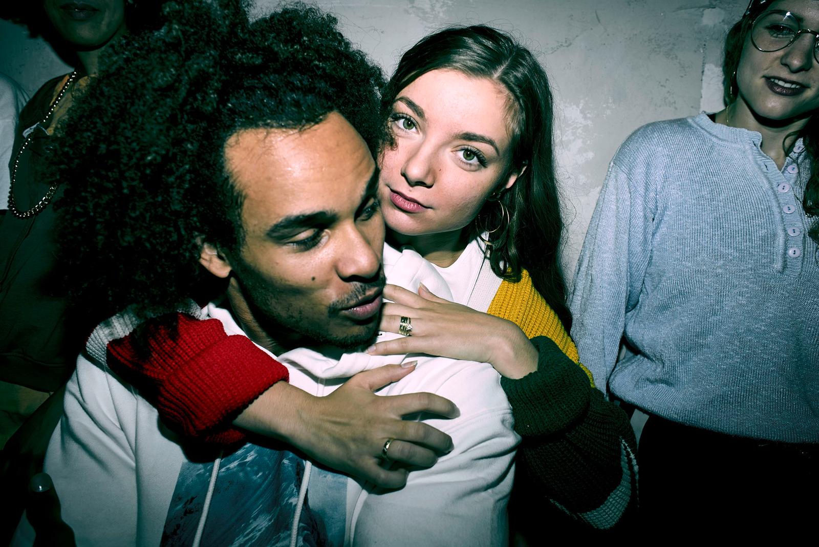 A young woman with curly hair and a man with a beard and mustache, both wearing casual clothing, embracing in a dimly lit room.