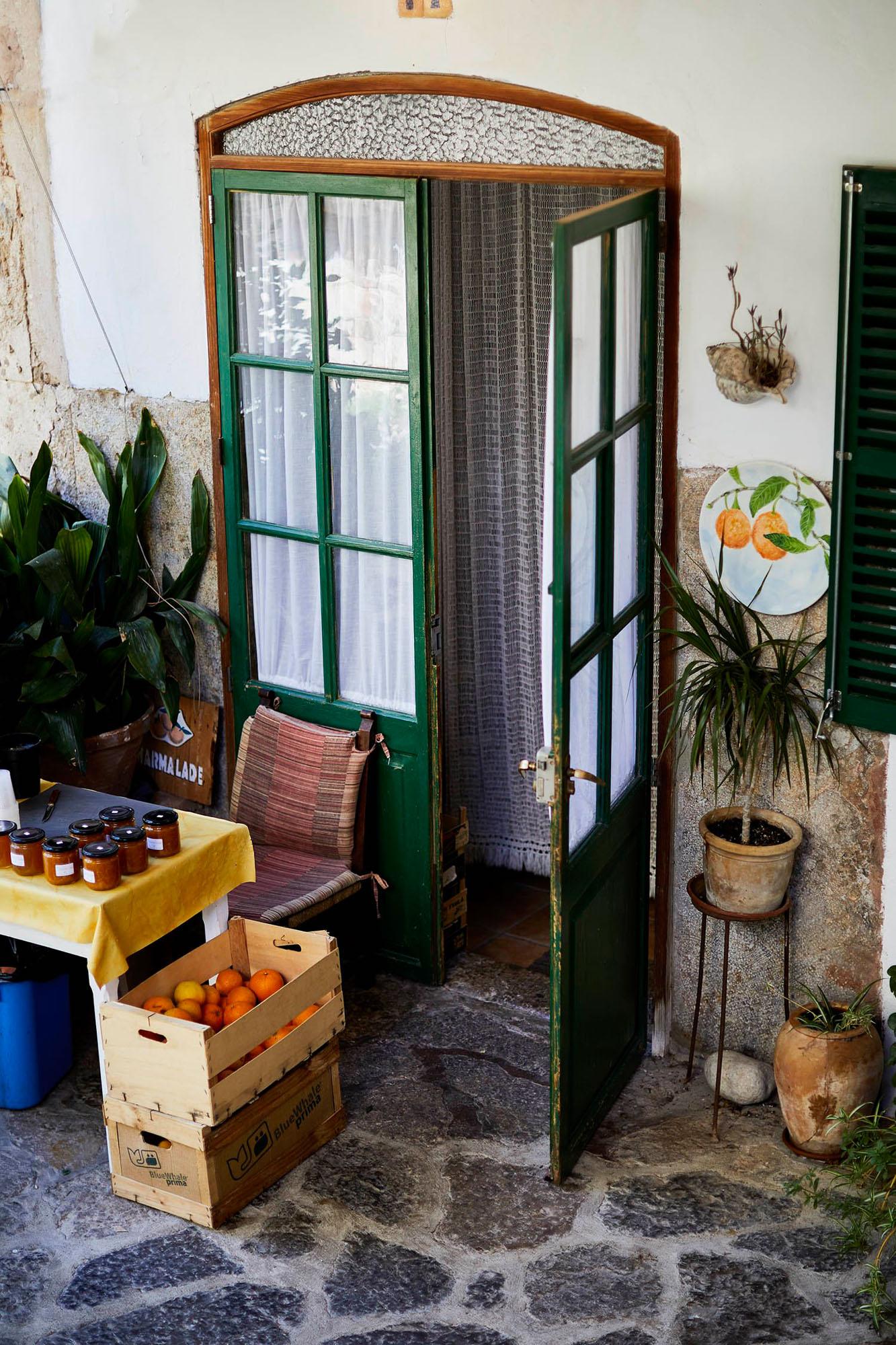 A small, rustic, outdoor market or shop, with a green door and a sign that says 'J' in a simple, hand-written font, surrounded by potted plants and a few crates of fresh produce.