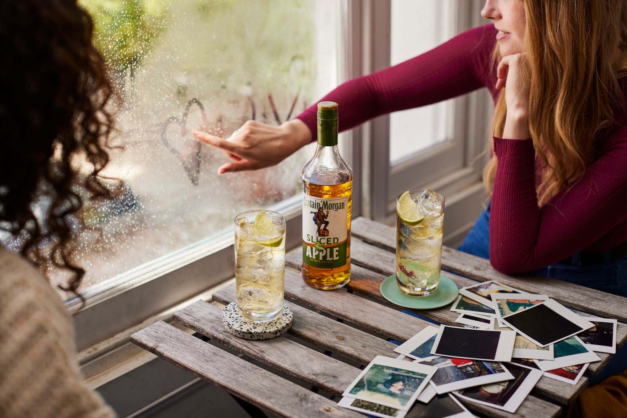 Two women sit at a wooden table by a window, with a bottle of Captain Morgan Sliced Apple rum and two glasses of rum and soda in front of them. The woman on the left is gesturing.
