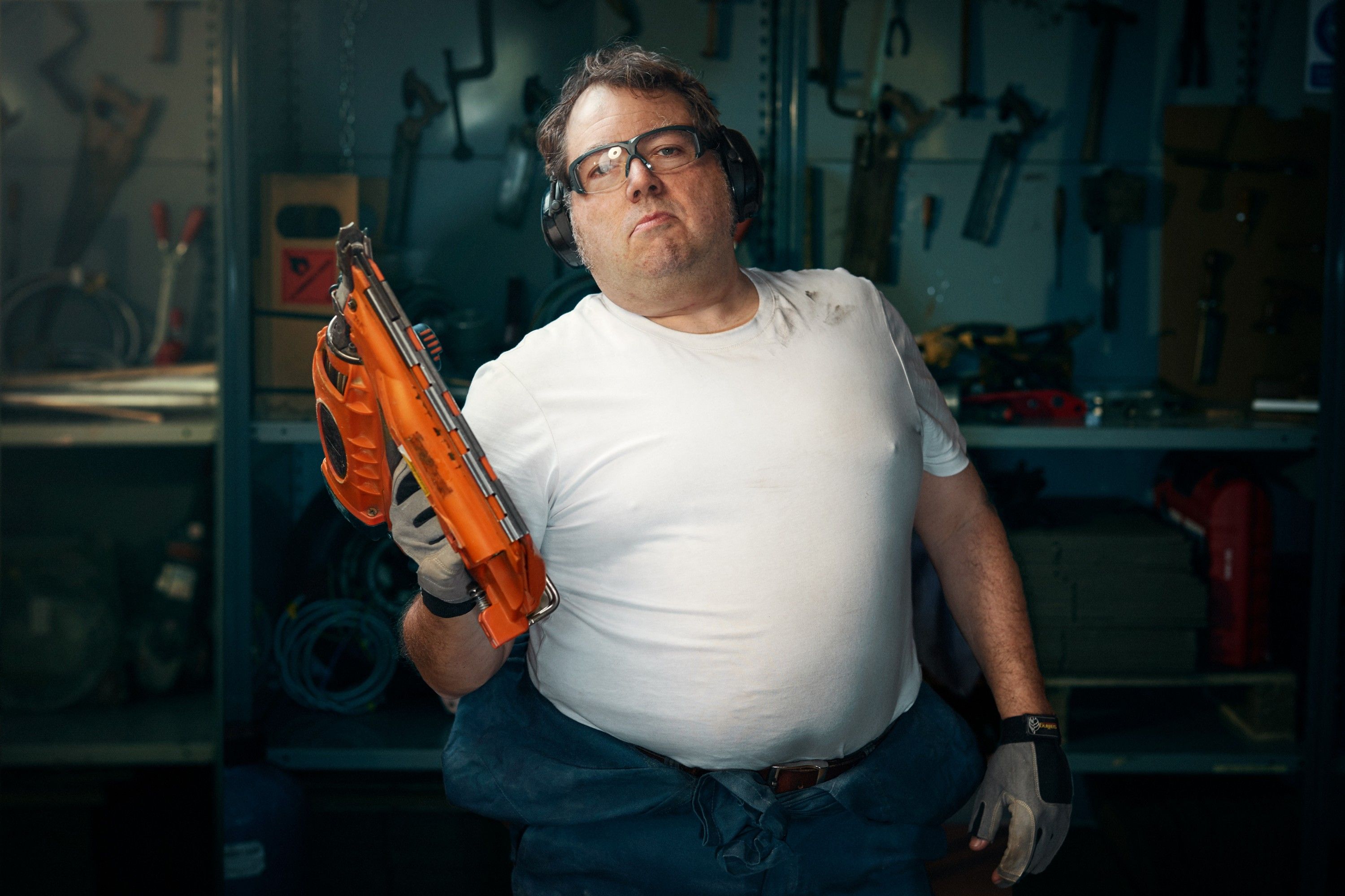A man in a white t-shirt, blue jeans, and safety goggles, holding an orange power tool in a workshop or garage.