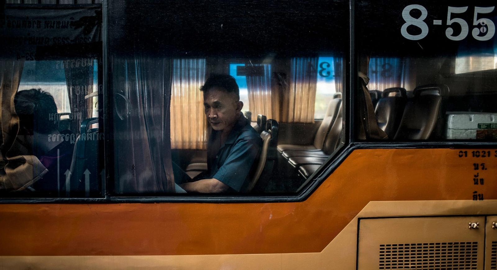 A man in a blue shirt is sitting in a bus, looking out the window at an urban cityscape.