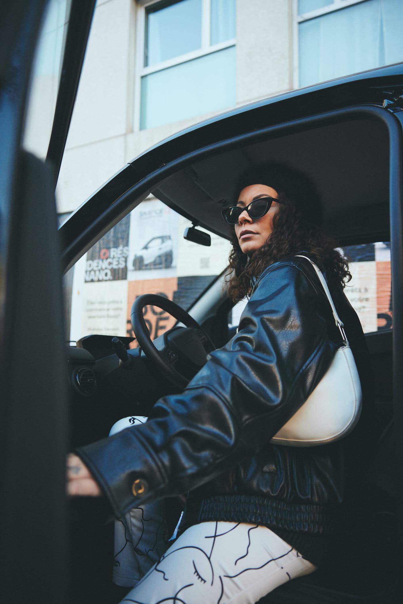 A woman with curly hair and a black leather jacket sits in the driver's seat of a car, wearing sunglasses and looking directly at the camera with a serious expression. The car's.
