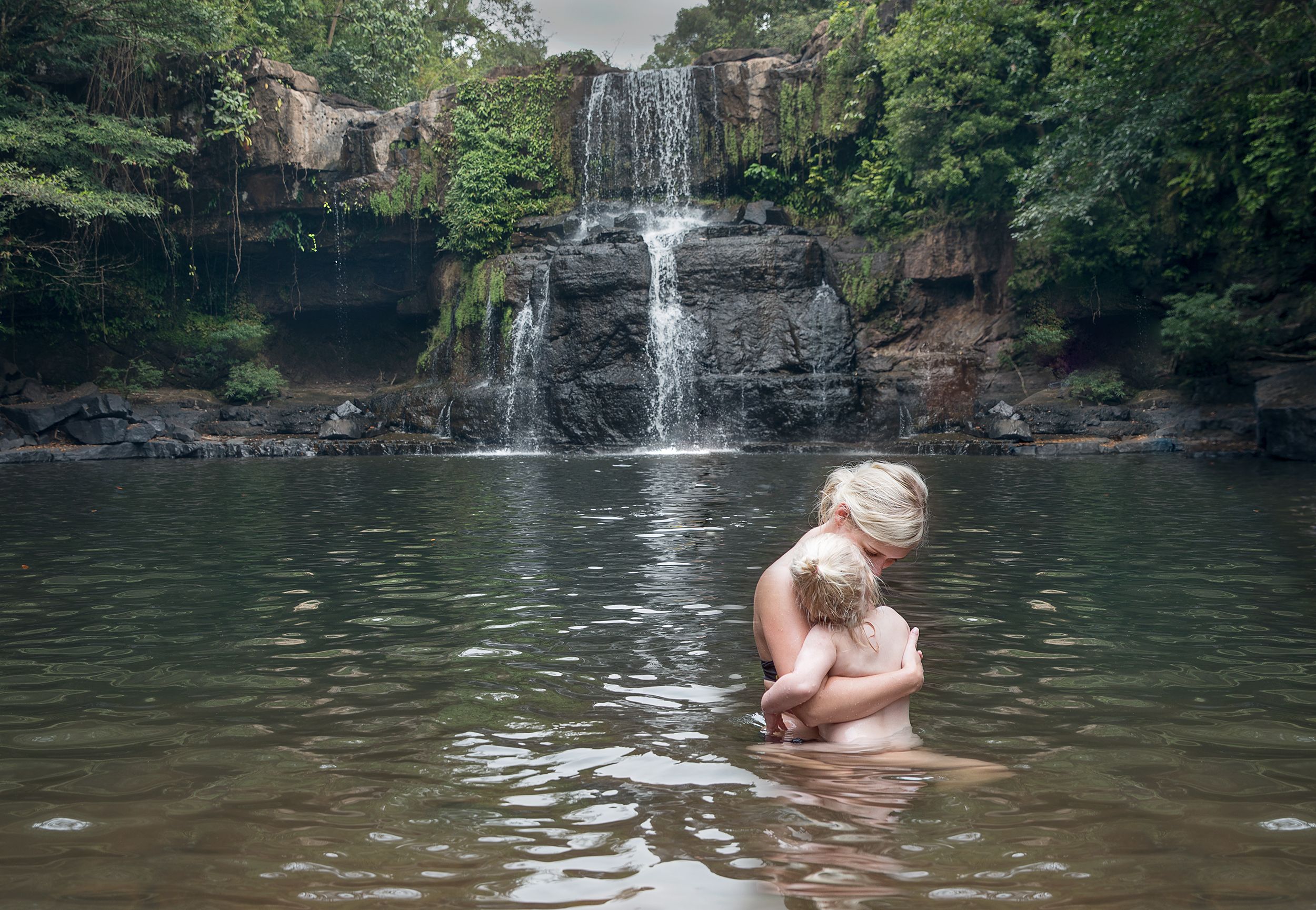 A woman in a black bikini swims with a young child in a natural, rocky, freshwater lake with a waterfall in the background.