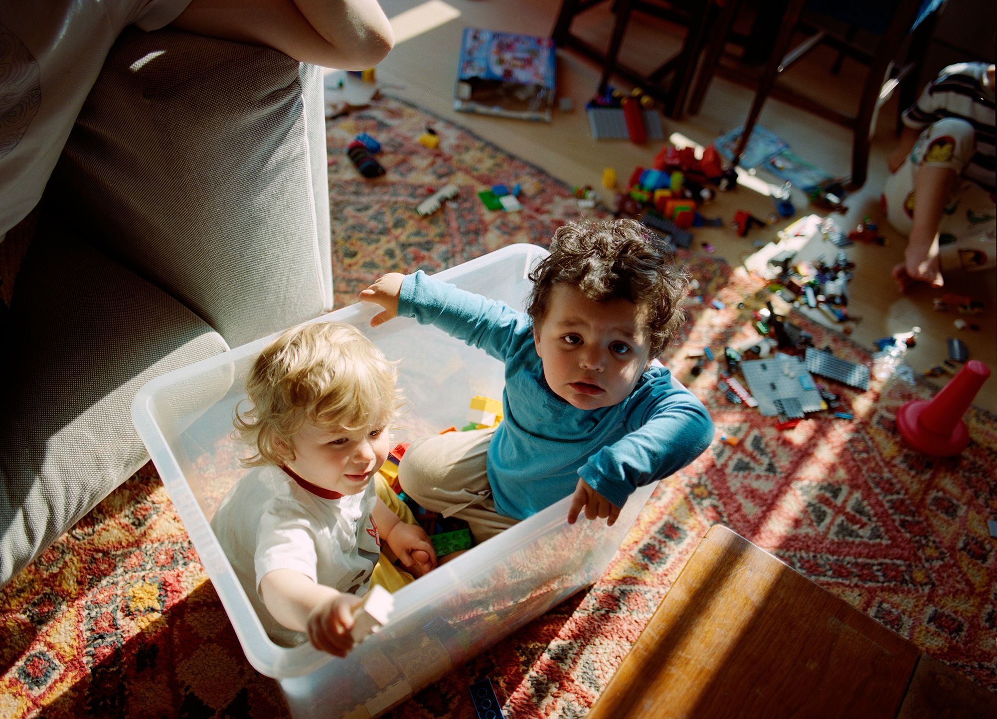 Two young children playing in a toy box filled with blocks and toys, surrounded by a messy room with toys and blocks scattered on the floor.