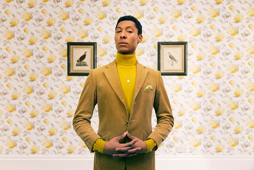 A young man in a tan suit and yellow turtleneck standing in front of a floral wallpaper with two bird prints on the wall.