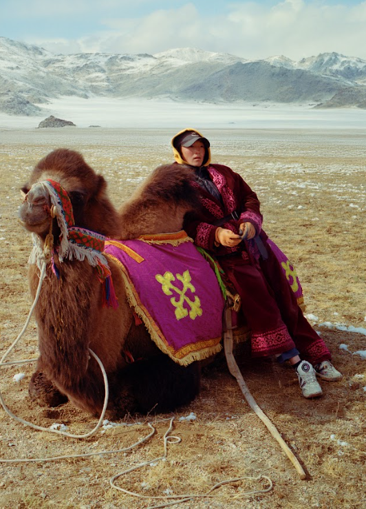 A person in traditional Mongolian attire sitting on a camel in a snowy mountain landscape.