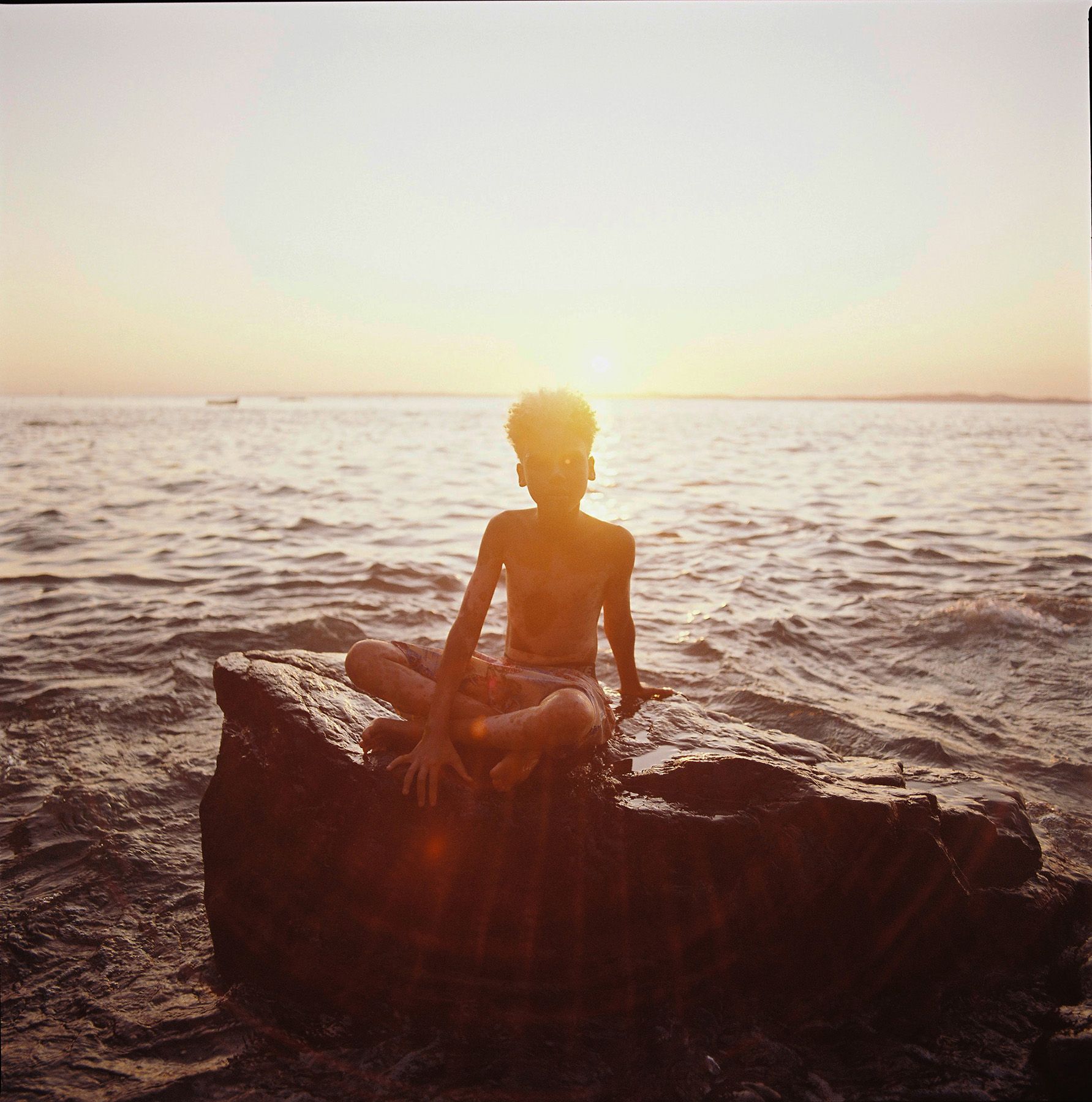 A young boy sitting on a rock at the beach, wearing a pair of shorts and a t-shirt, with the sun setting in the background.