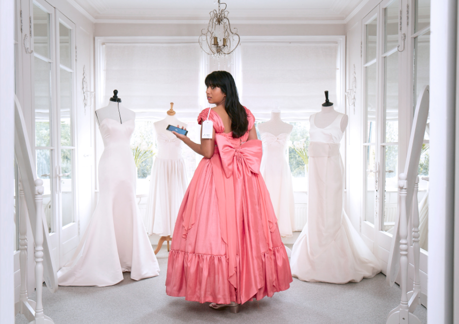 A woman in a pink ball gown holding a tablet in a room with several white wedding dresses on display.