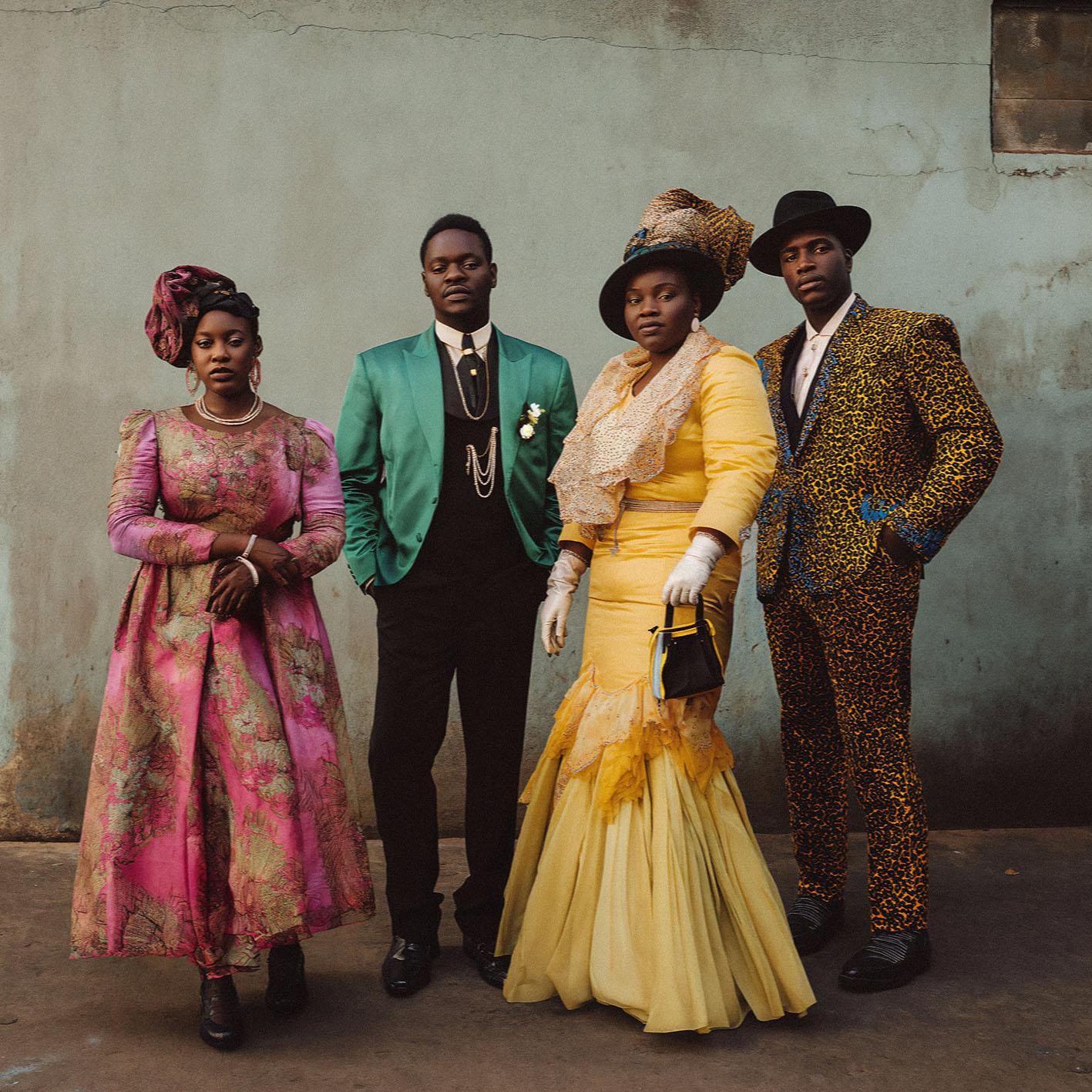 A group of four people, two men and two women, dressed in 19th-century Victorian-era clothing, standing in front of a plain background.