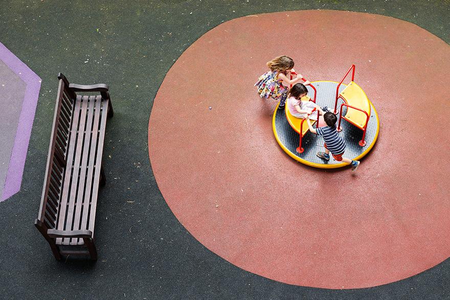 Aerial view of a playground with a yellow and red merry-go-round with three children playing on it, surrounded by a bench and a playground area.