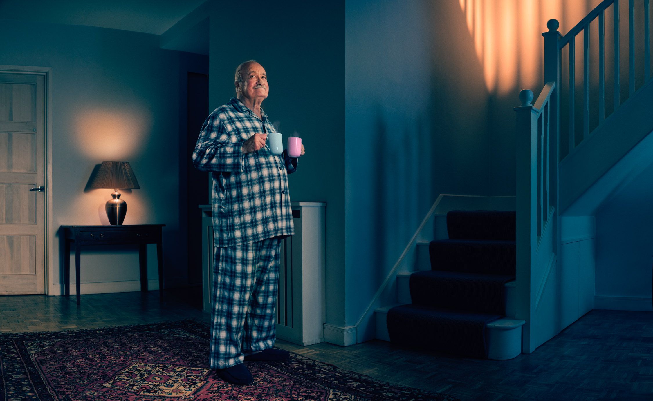 A man in plaid pajamas holds two mugs in a dimly lit hallway with a rug and a table lamp.