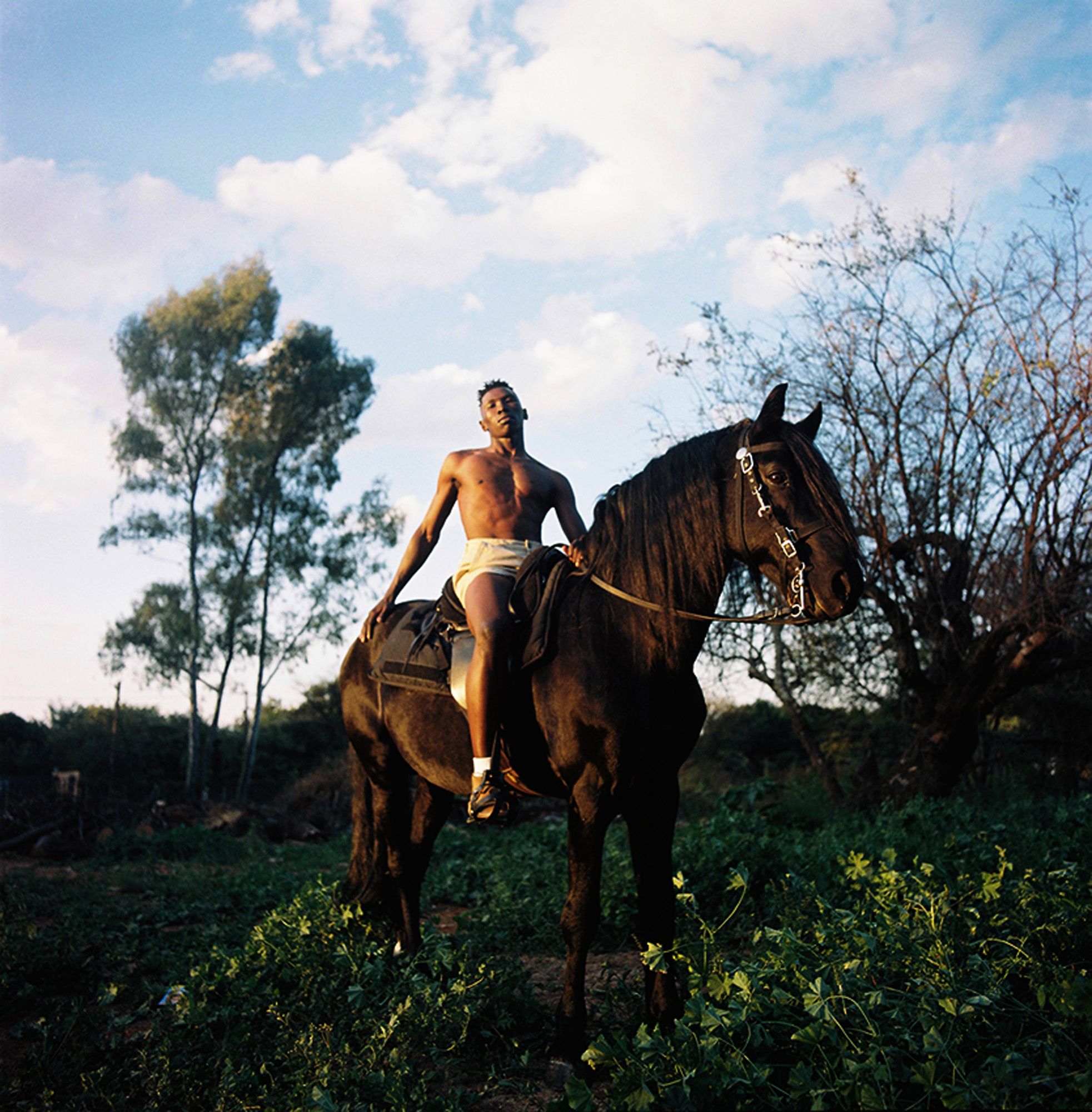 A man in a pair of white underwear is riding a black horse in a field with trees and a blue sky in the background.
