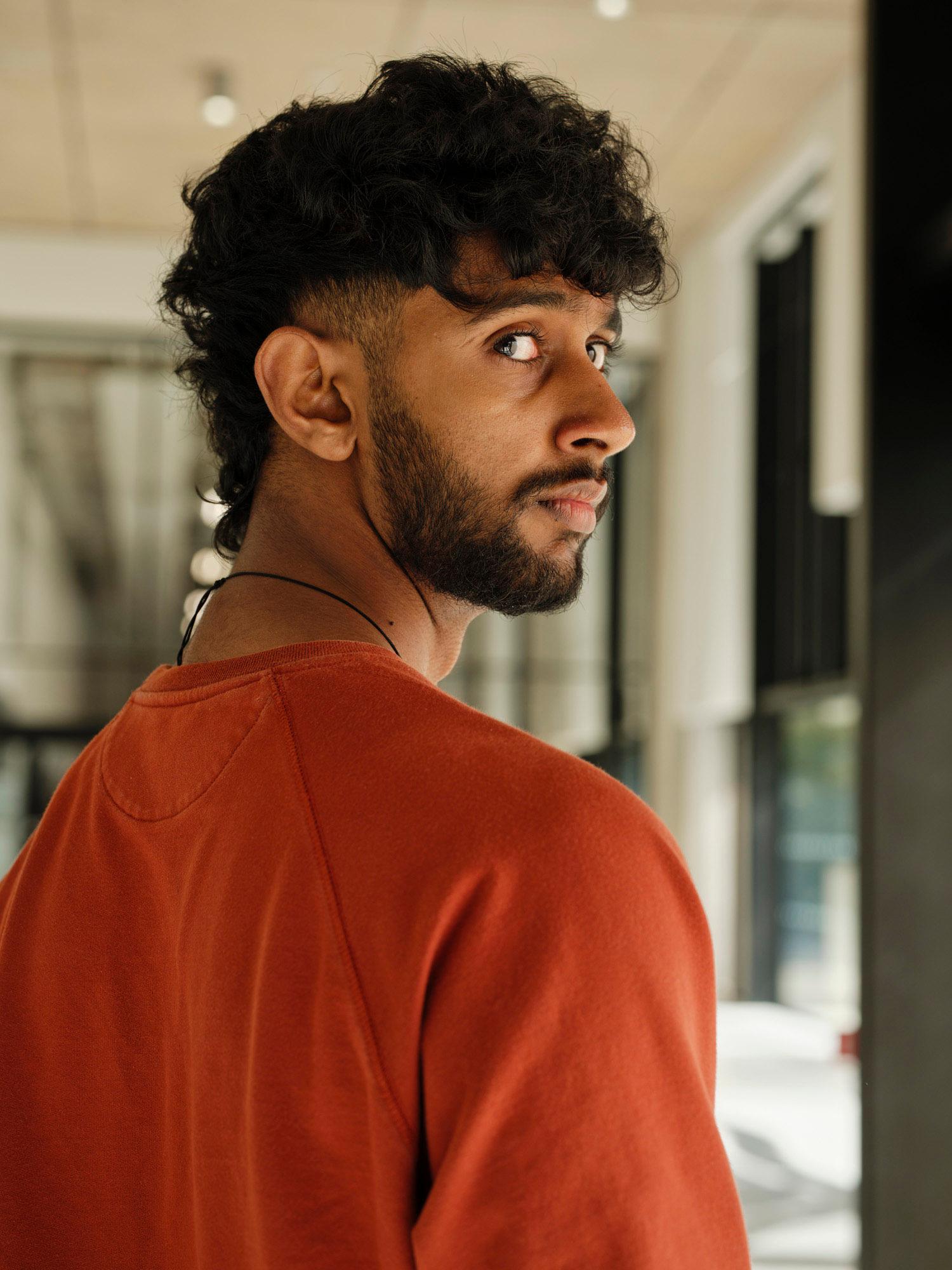 A young man with dark hair and a beard, wearing an orange t-shirt, looking over his shoulder with a curious expression.