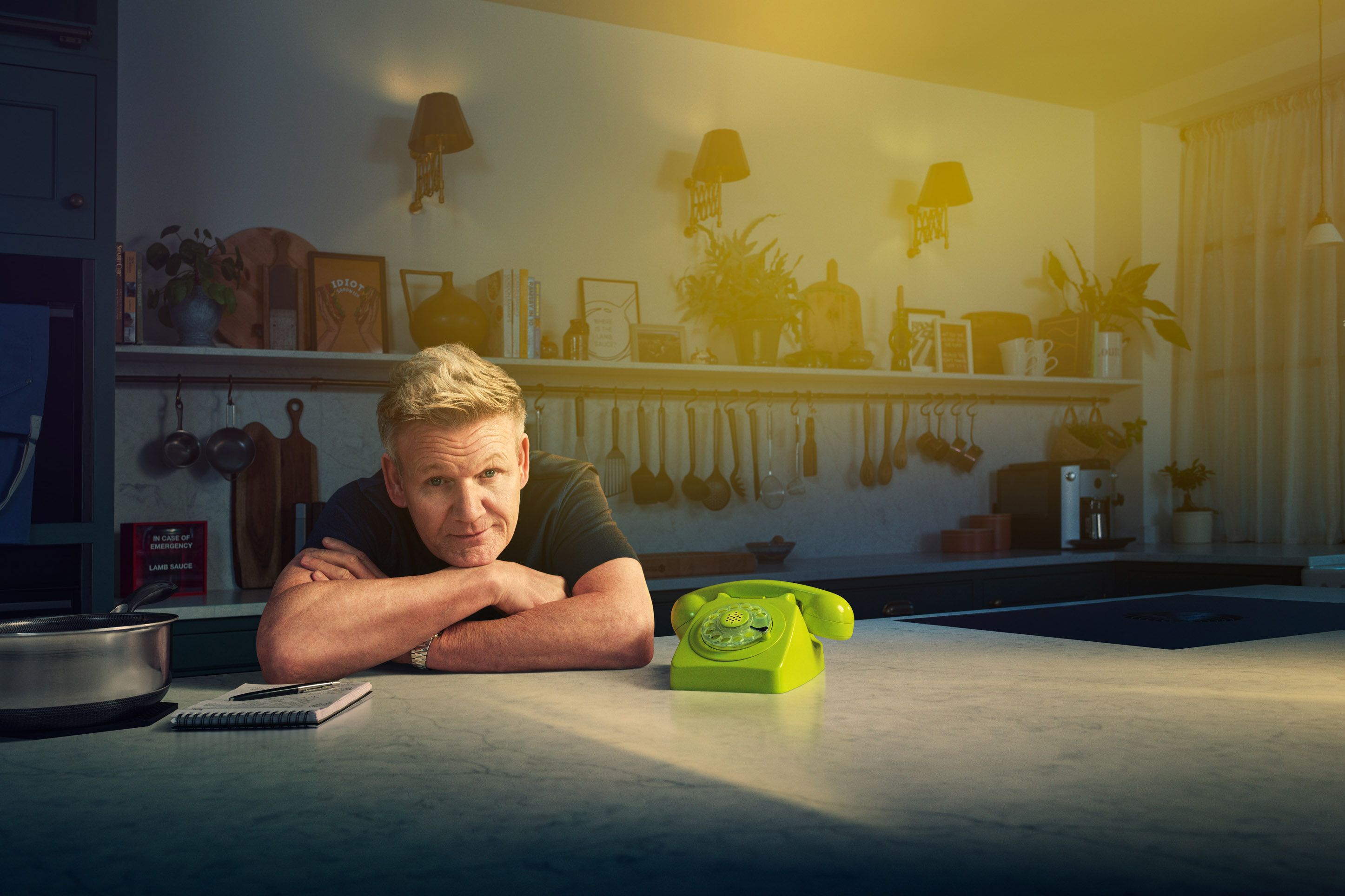The image shows Gordon Ramsay leaning on a kitchen island for a PolyAI campaign photographed by Samuel McElwee. He is looking directly at the camera with a slight smile while resting his chin on his crossed arms. Beside him sits a bright lime-green rotary phone, contrasting with the warm, dimly lit kitchen background that features shelves of cookbooks and plants. A dramatic yellow light flare glows from the upper right corner of the frame.