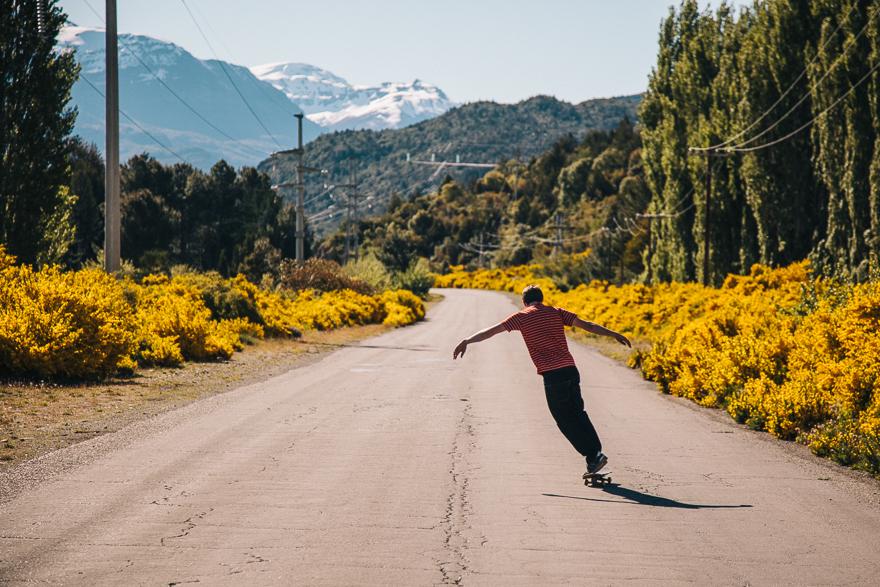A person skateboards down a long, straight road through a mountainous landscape wearing a red and white striped shirt and black pants.